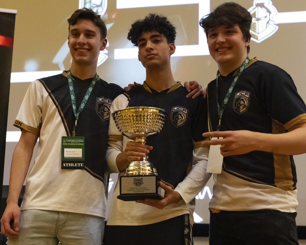 University of Central Florida Rocket League players wearing grey, gold, and white jerseys pose with trophy.