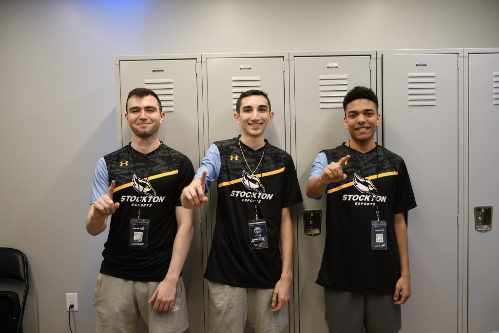 Stockton University Rocket League players wearing grey jerseys pose in front of lockers making the "L" shape with their hands